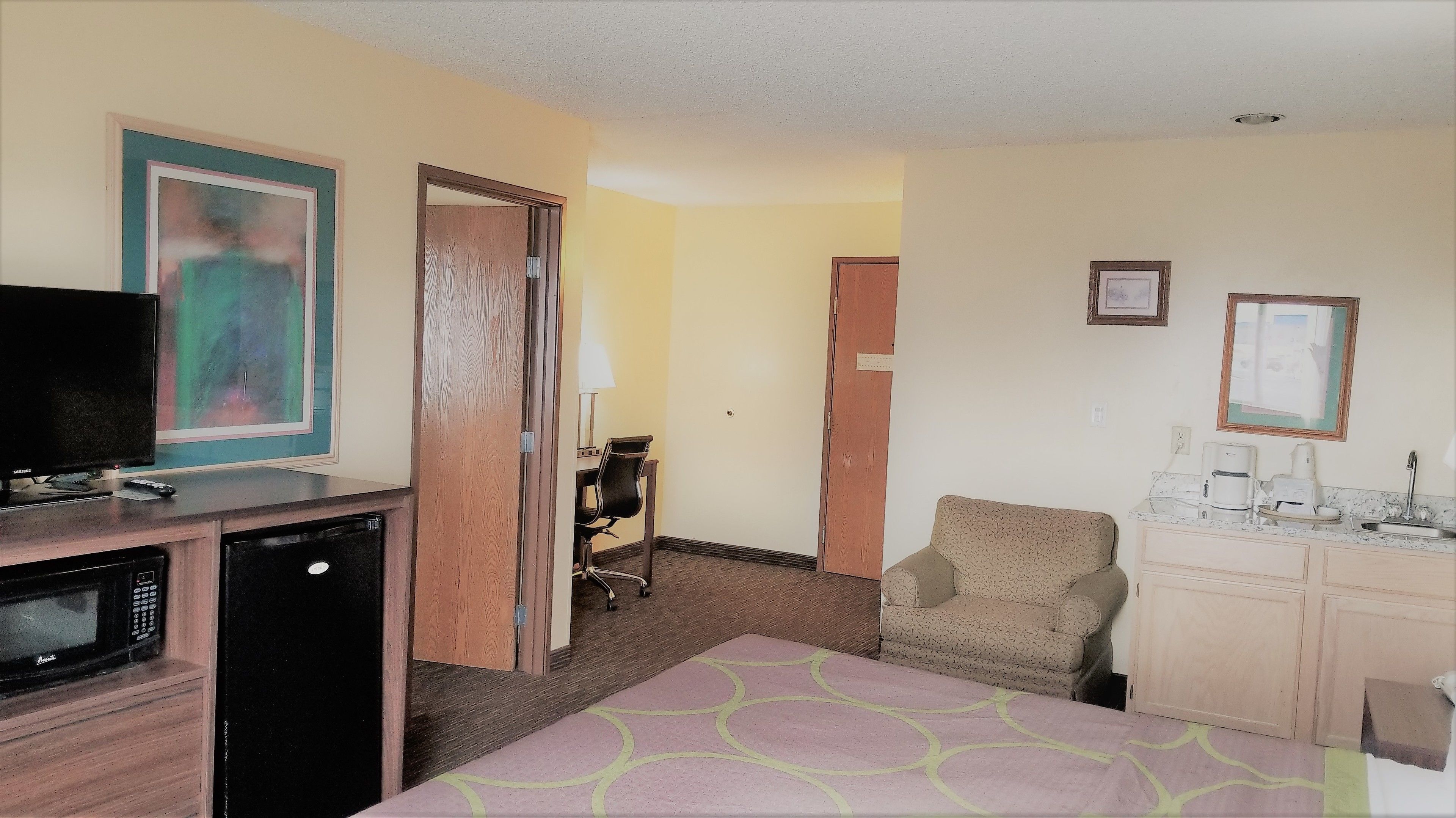 A hotel room with a bed, a desk with a computer, a television, a microwave, a chair, and a kitchenette with a coffee maker and a sink.