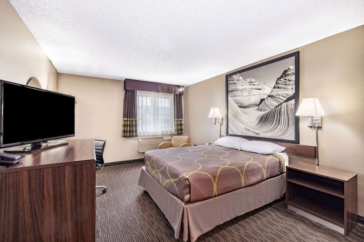 A hotel room with a bed, a desk with a TV, a chair, a window with curtains, and a wall-mounted lamp.