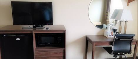 A hotel room with a flat-screen TV, a microwave, a refrigerator, a desk with a chair, a lamp, and a mirror.