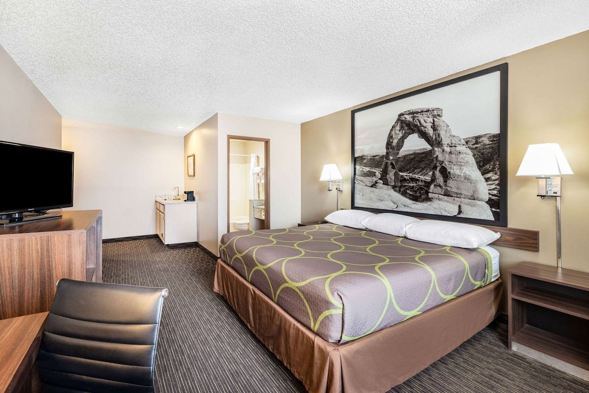 A hotel room with a bed, a desk with a television, a chair, a bathroom, and a large framed picture on the wall.