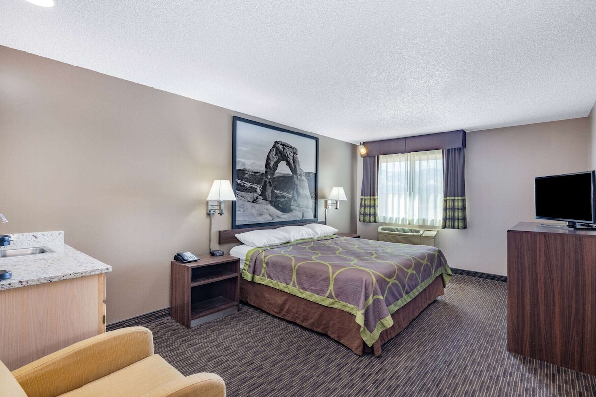 A hotel room with a bed, a desk, a TV, a chair, a window with curtains, and a painting on the wall.