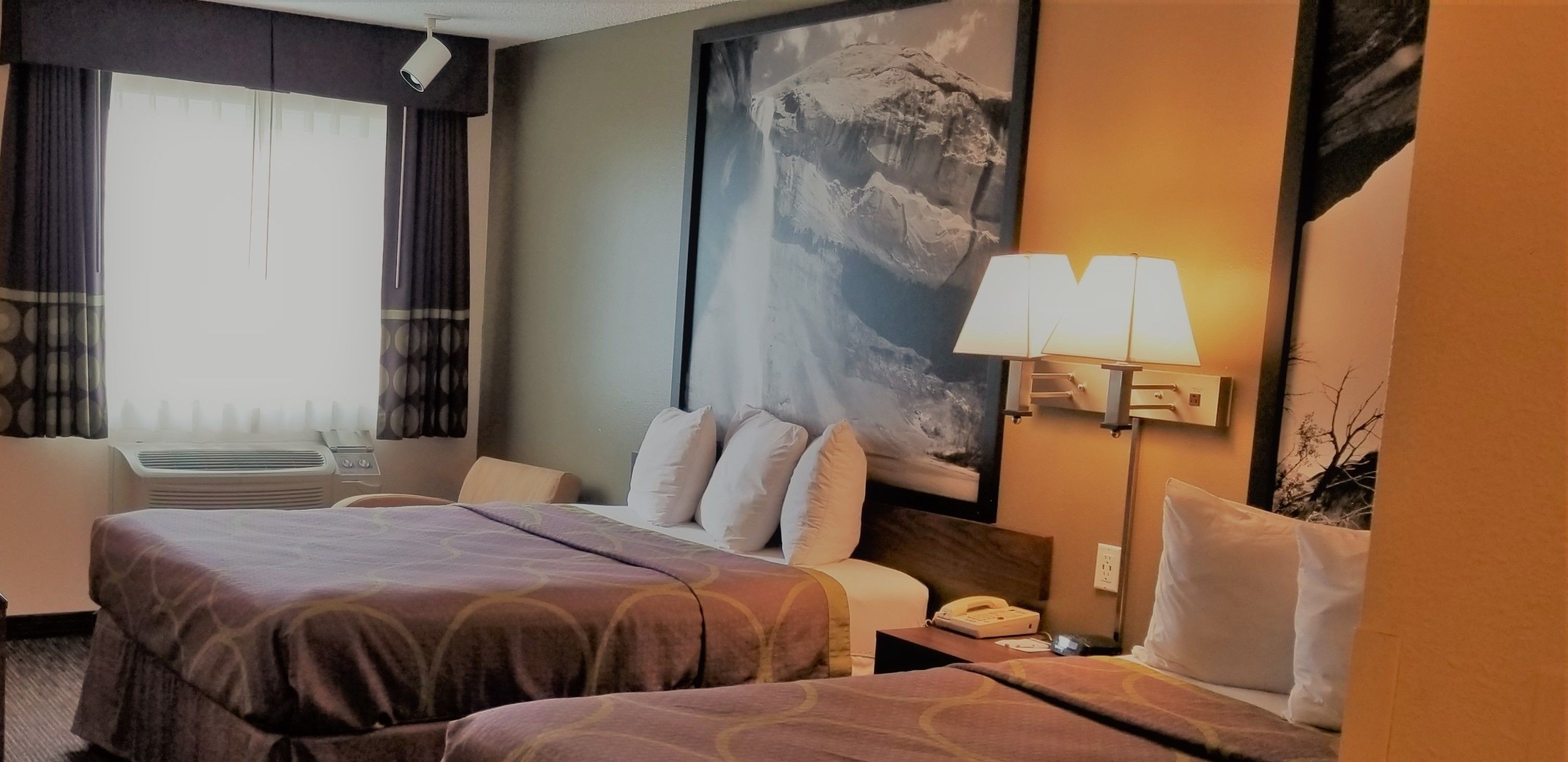 A hotel room with two beds, a large window with curtains, a mounted painting, a desk with a lamp, and a telephone.