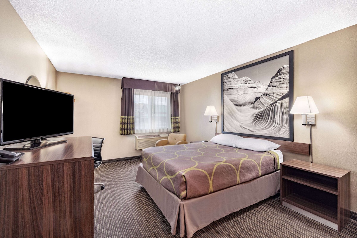 A hotel room with a bed, a desk with a TV, a chair, a window with curtains, and a painting on the wall.