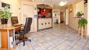 A reception area with a desk, computer, and office chair. There is a reception desk with a red counter and a door in the background. The floor is tiled, and there are potted plants on the right side. - Motel 6 Wilsonville, OR - Portland (Wilsonville)