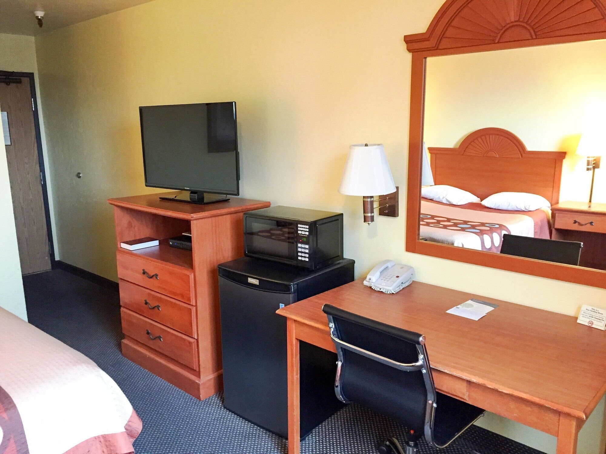 A hotel room with a wooden desk, a television, a microwave, a telephone, and a mirror reflecting a bed with pillows.