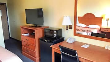 A hotel room with a wooden desk, a television, a microwave, a telephone, and a mirror reflecting a bed with pillows.