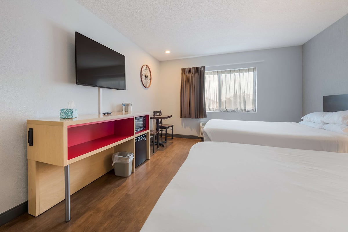 A hotel room with two beds, a TV mounted on the wall, a desk with a chair, and a window with curtains.