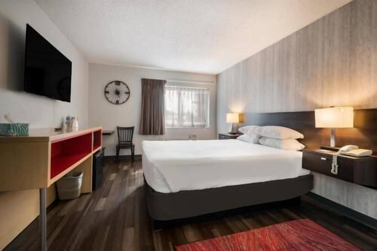 A hotel room with a bed, a TV mounted on the wall, a desk with a chair, and a window with curtains.