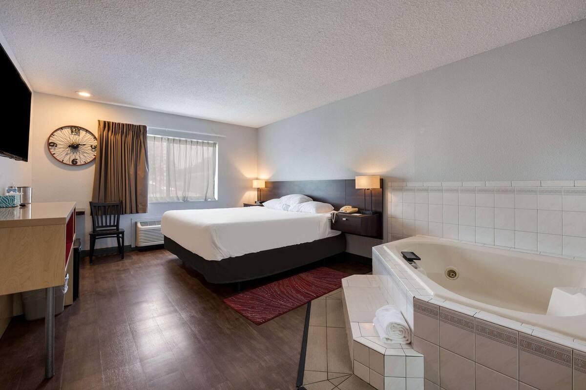 A hotel room with a bed, a desk, a chair, a large clock, a window with curtains, a bathroom with a bathtub, and a towel rack.