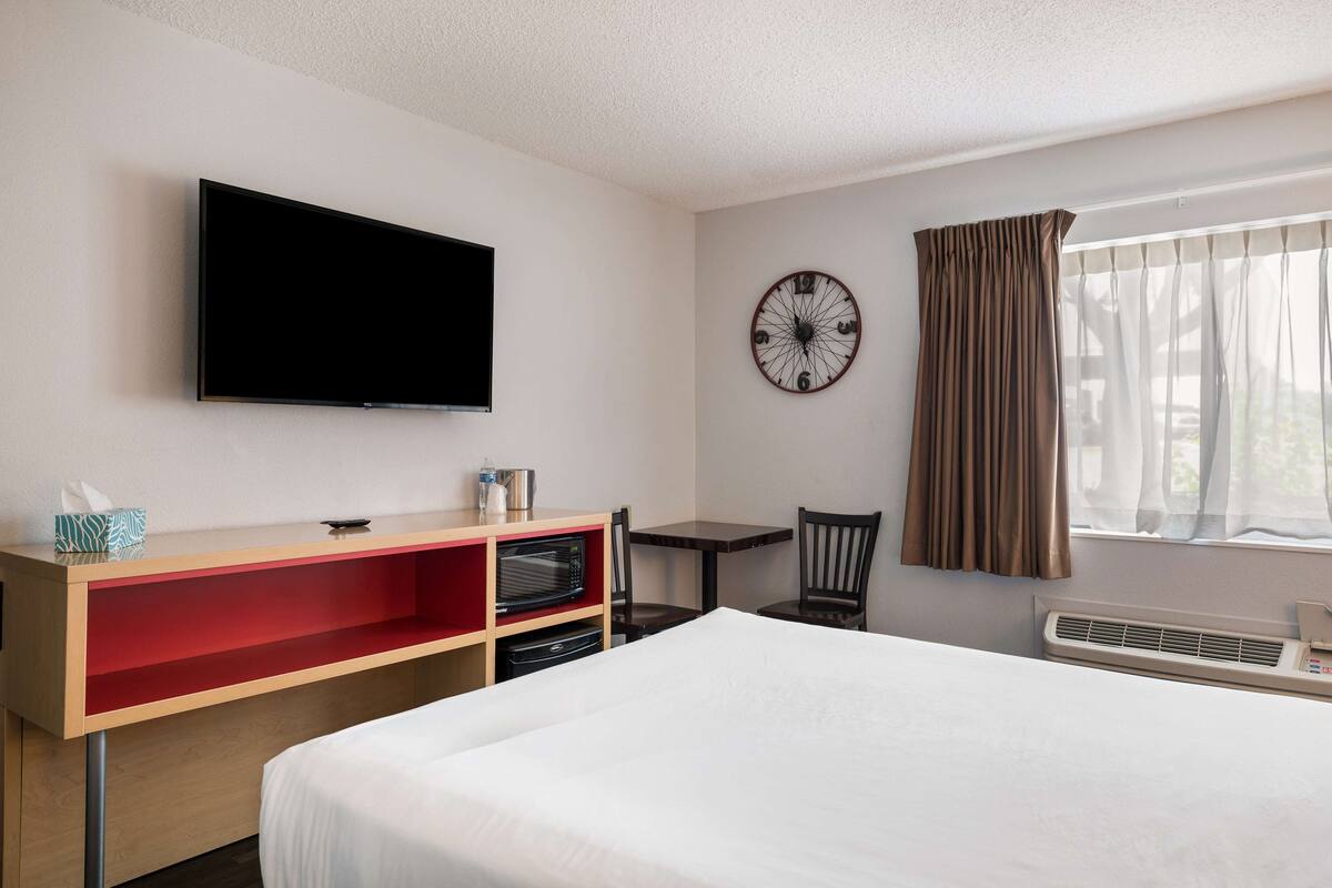 A hotel room with a bed, a TV mounted on the wall, a clock, a window with curtains, a desk with a microwave, and a chair.