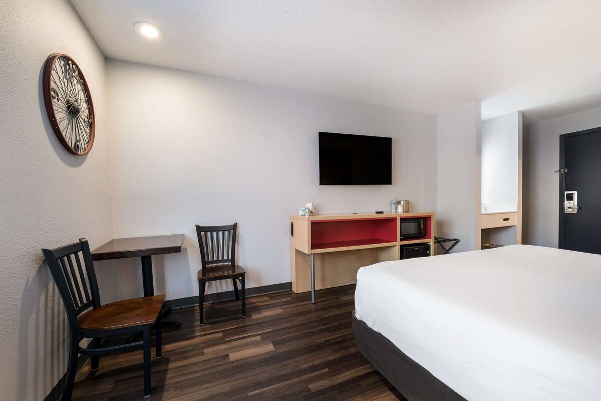 A hotel room with a bed, a desk with a chair, a television, and a wall-mounted clock.