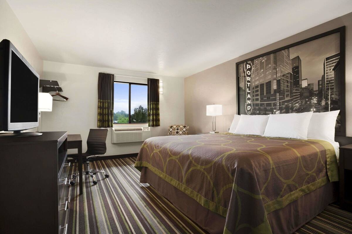 Une chambre d’hôtel avec un lit, un bureau, une chaise, une télévision, une fenêtre avec des rideaux et un tableau représentant un paysage urbain accroché au mur.