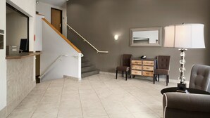 Un vestíbulo de hotel con mostrador de recepción, una escalera, una zona de estar con sillas, una lámpara y un espejo en la pared.