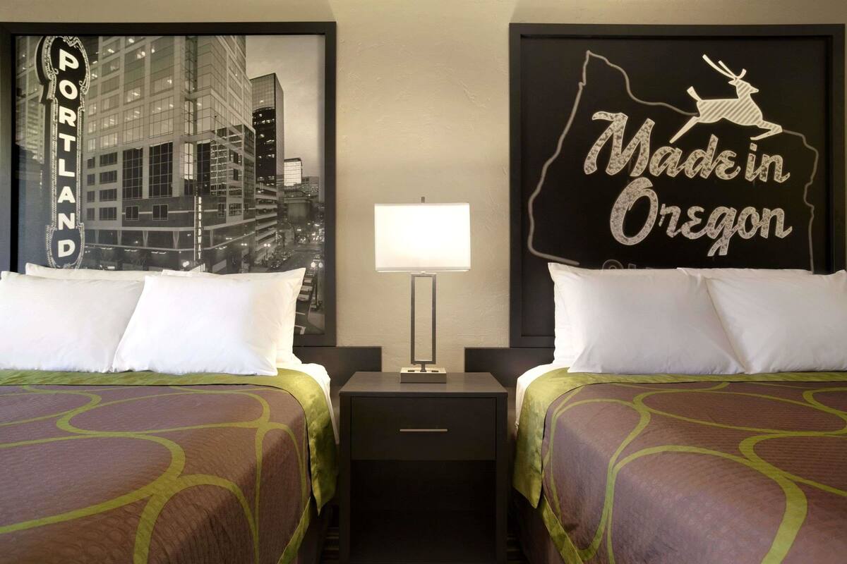 Une chambre d’hôtel avec deux lits, une table de chevet avec une lampe et des œuvres d’art murales représentant Portland et l’Oregon.
