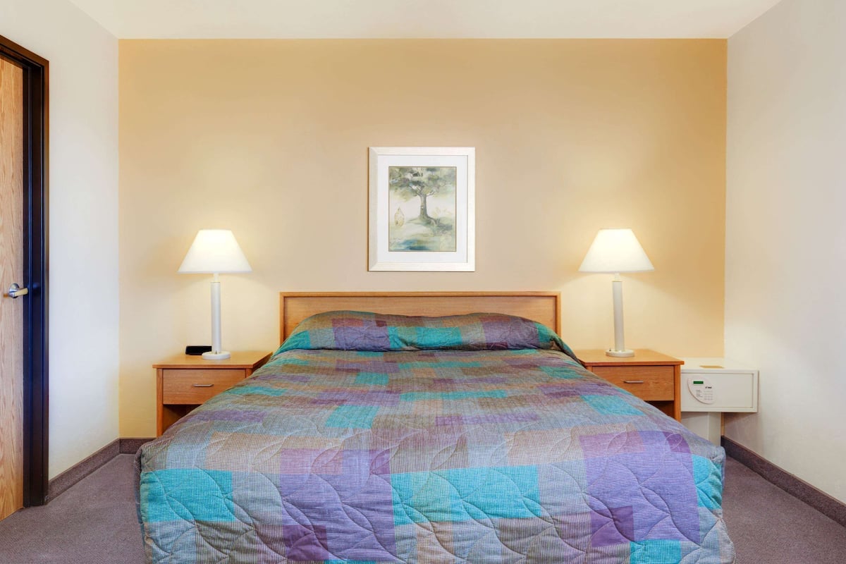 A neatly made bed with a colorful quilt, two bedside lamps, a nightstand, and a framed picture on the wall.