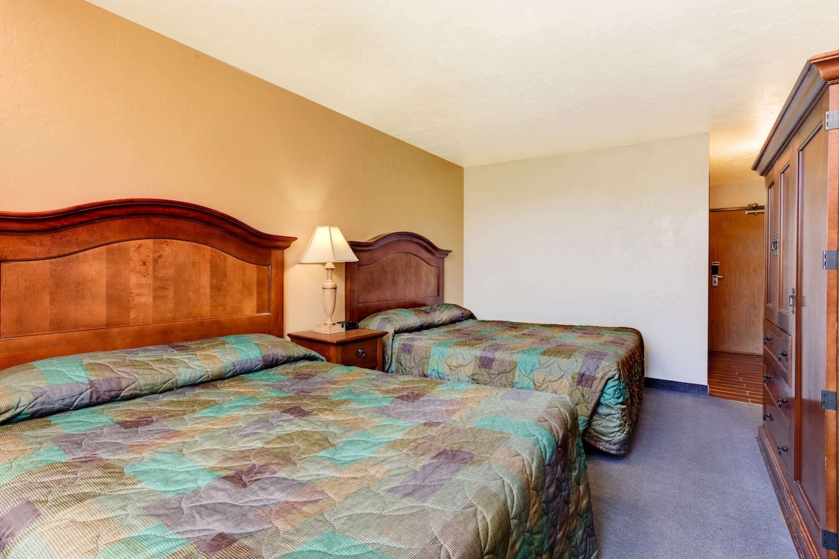 A hotel room with two beds, a wooden headboard, a nightstand with a lamp, and a wardrobe.
