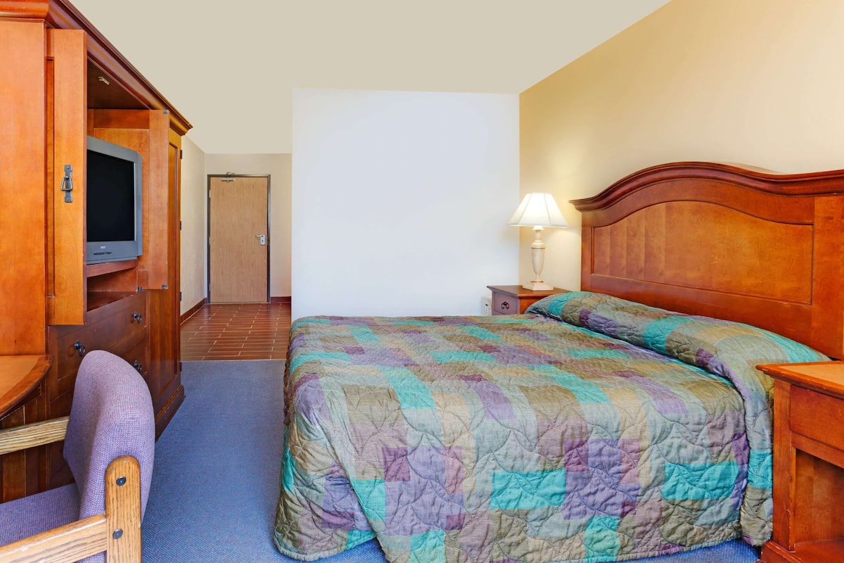 A hotel room with a bed, a desk with a television, a chair, and a nightstand with a lamp.