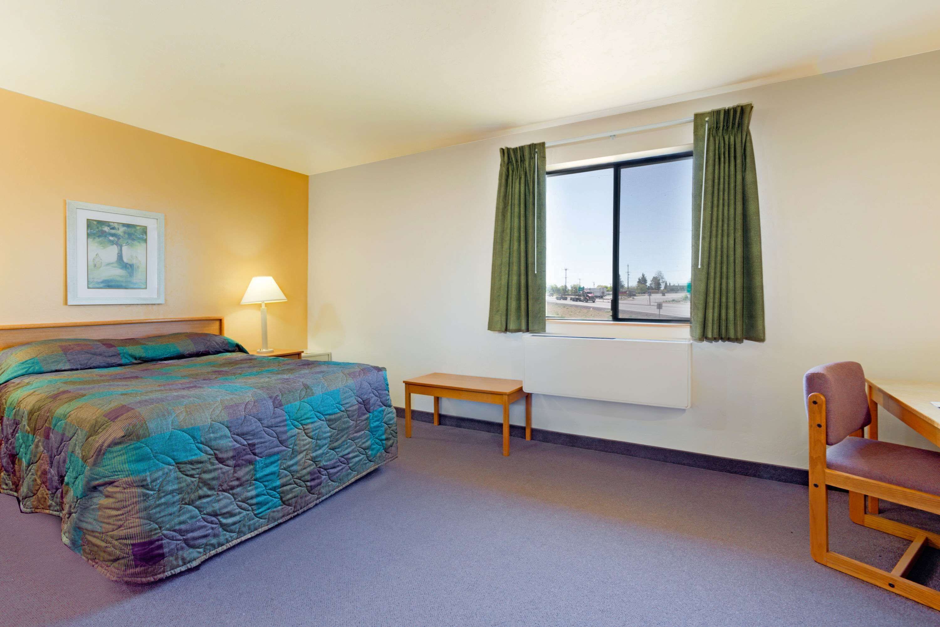 A hotel room with a bed, a small table, a chair, a window with curtains, and a painting on the wall.