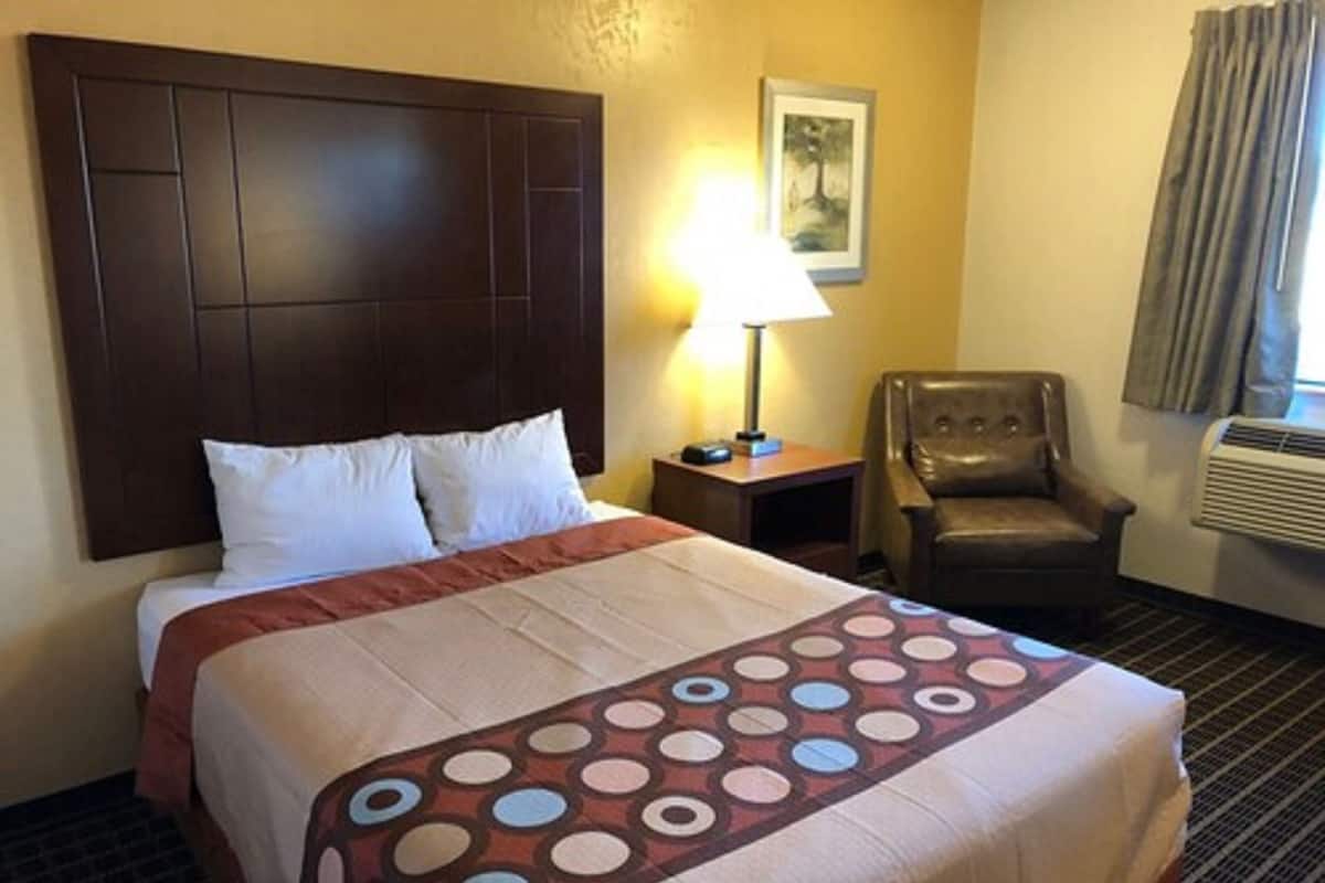 A hotel room with a large bed, a chair, a nightstand, a lamp, and an air conditioning unit.