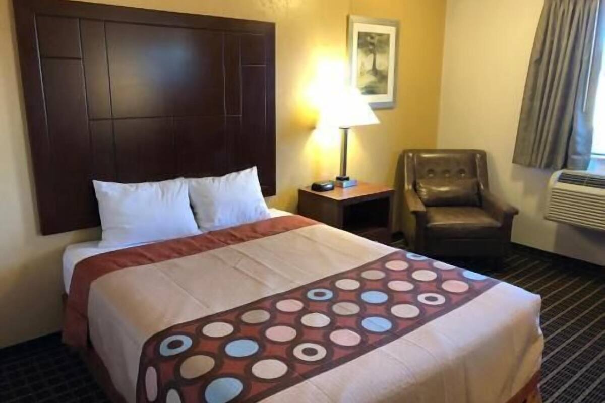 A hotel room with a large bed, a chair, a nightstand, a lamp, and an air conditioning unit.