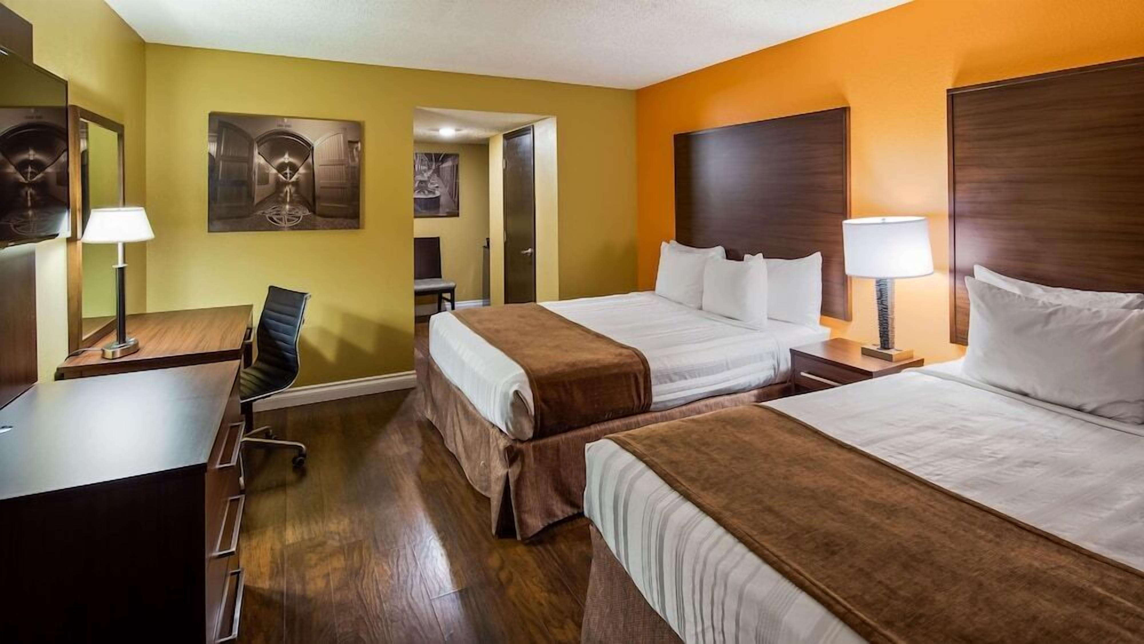Quarto de hotel com duas camas, uma escrivaninha, uma cadeira e uma mesa de cabeceira com abajur.