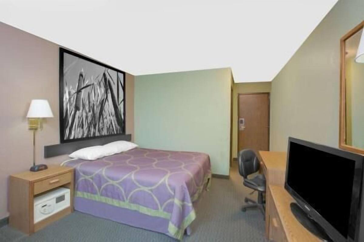 A hotel room with a bed, a desk, a television, a nightstand, and a painting on the wall.