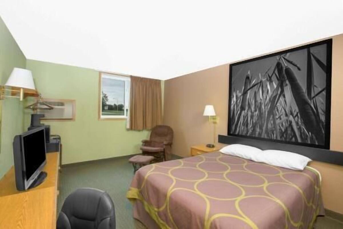 A hotel room with a bed, a desk with a computer, a chair, a window, and a painting on the wall.