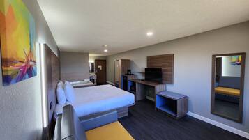 A modern hotel room with a large bed, a desk with a flat-screen TV, a mirror, and a small bathroom.