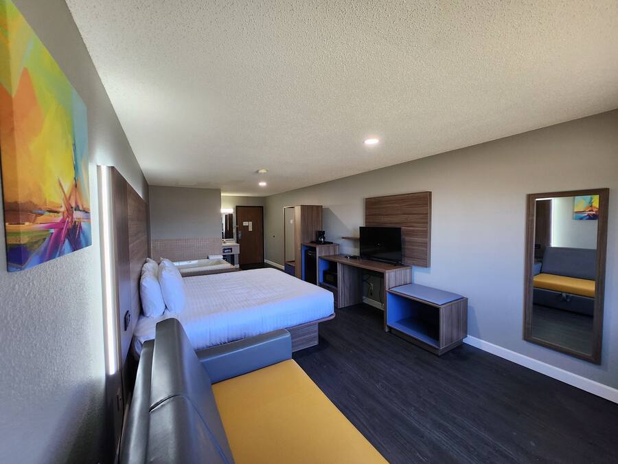 A modern hotel room with a large bed, a desk with a flat-screen TV, a mirror, and a small bathroom.