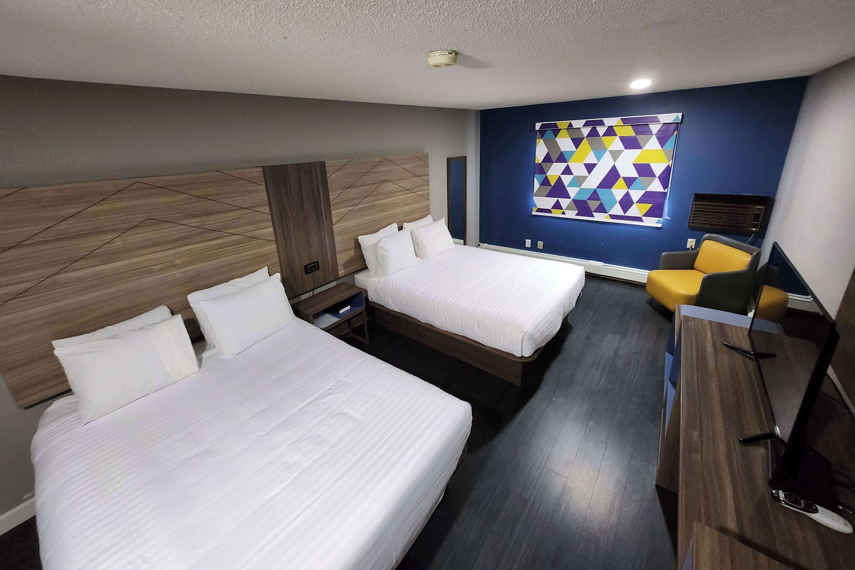 Habitación de hotel con dos camas, cabecera de madera, cuadro de arte geométrico y colorido en la pared, escritorio con silla y diseño moderno.