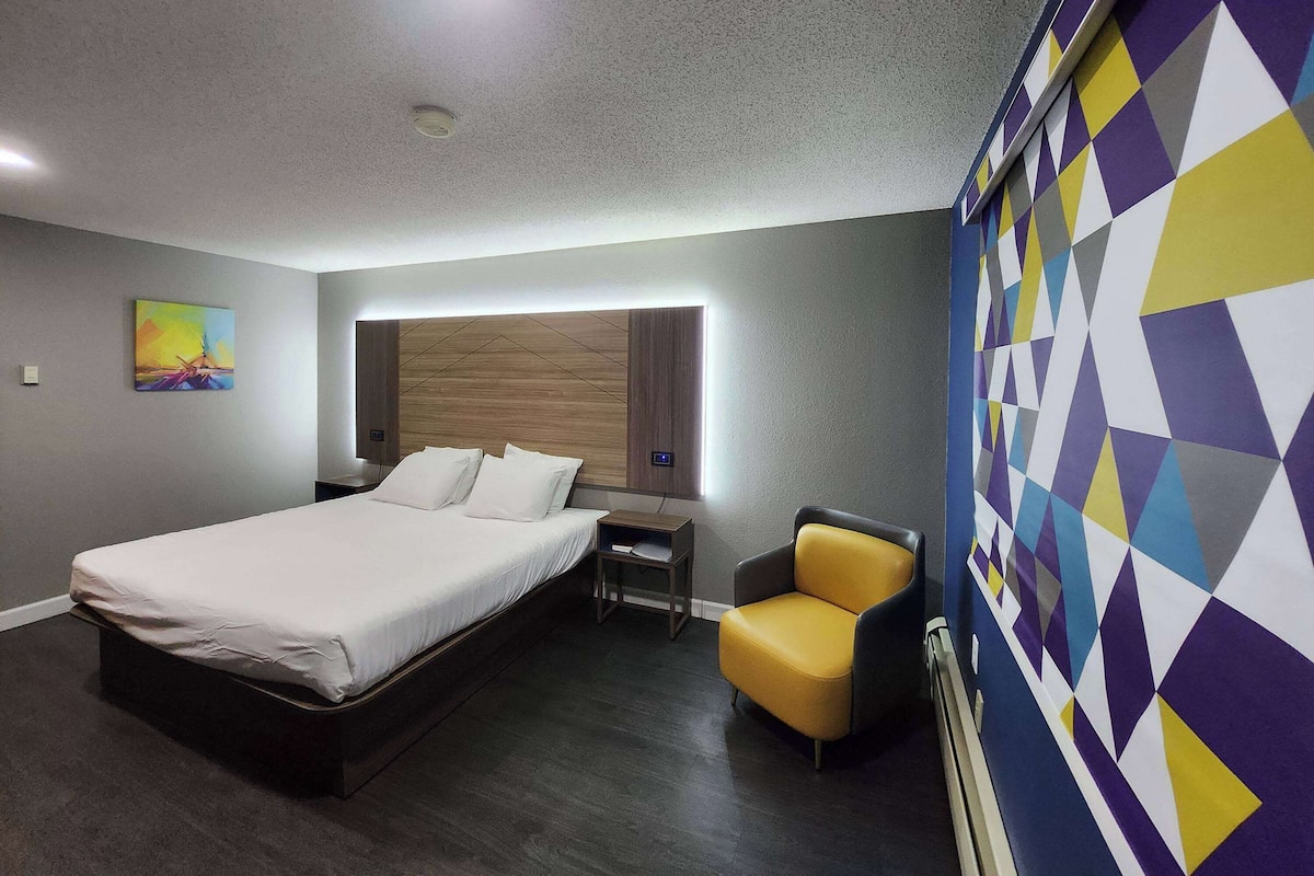 A modern hotel room with a large bed, a bedside table, a chair, and a colorful geometric-patterned wall.