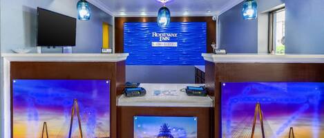 A hotel reception area with a large screen displaying the hotel name, a TV, and a reception desk with a digital display.