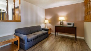 A room with a wooden floor, a gray sofa, a small table, a lamp, and a microwave on a cabinet. - Super 8 by Wyndham Pendleton (Pendleton)