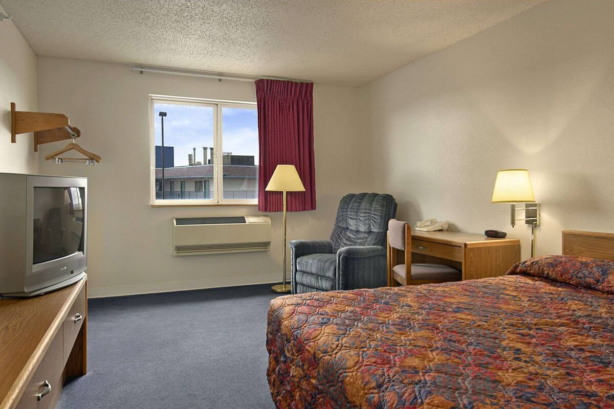 A hotel room with a bed, a desk, a chair, a TV, a window with a view of buildings, and a red curtain.