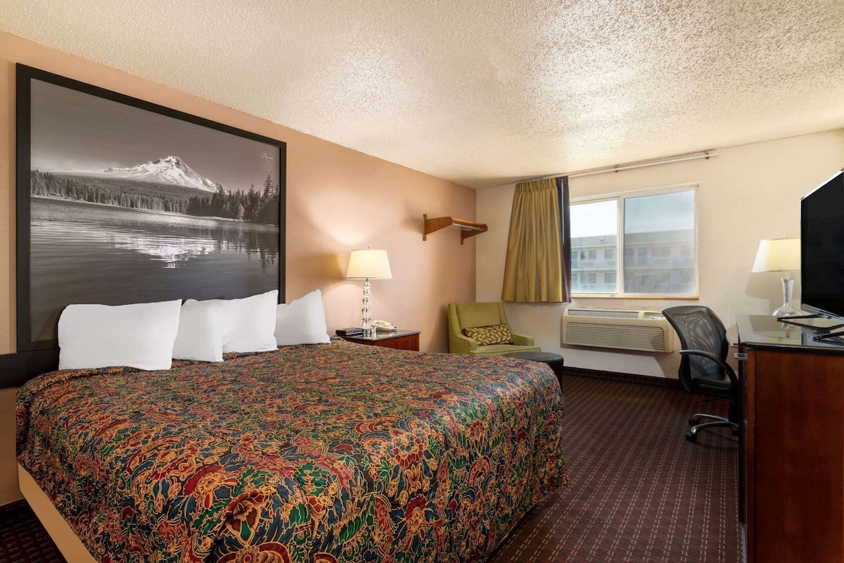 A hotel room with a large bed, a desk, a chair, a television, a window with curtains, and a wall-mounted air conditioning unit.