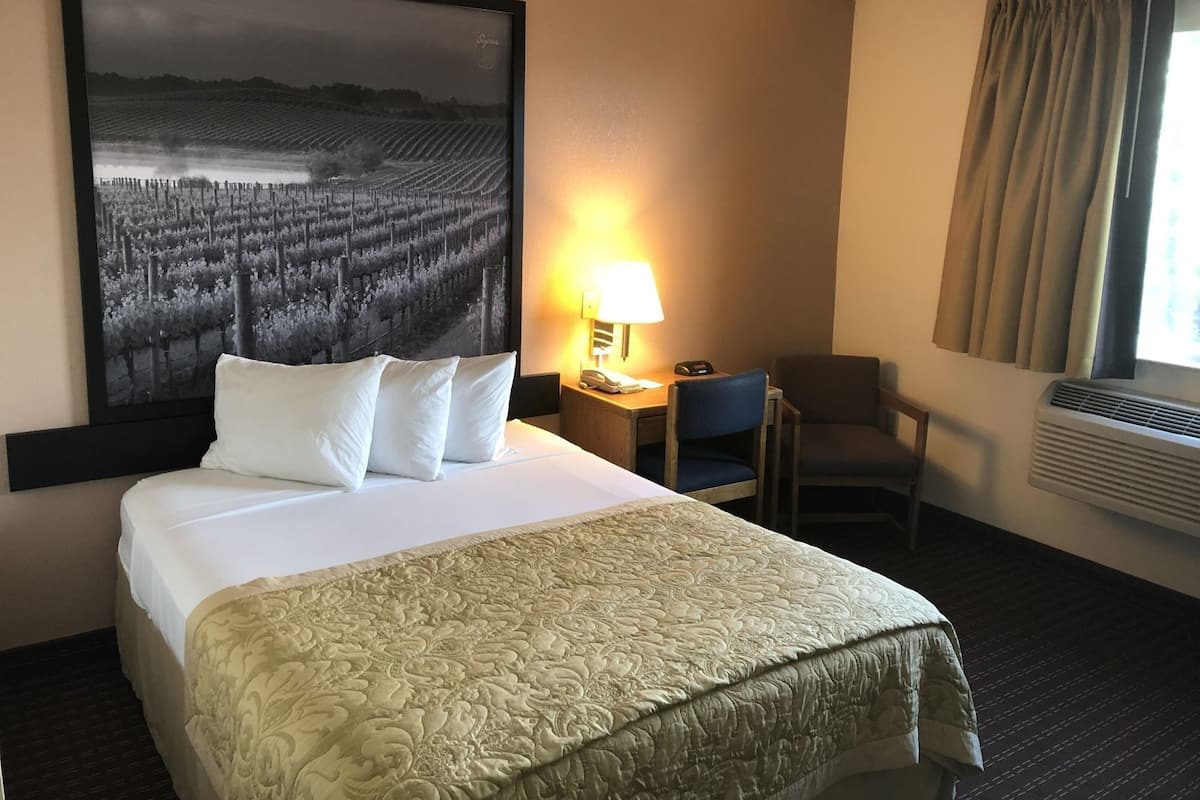 A hotel room with a large bed, a desk with a lamp, a chair, and a large framed picture of a vineyard.