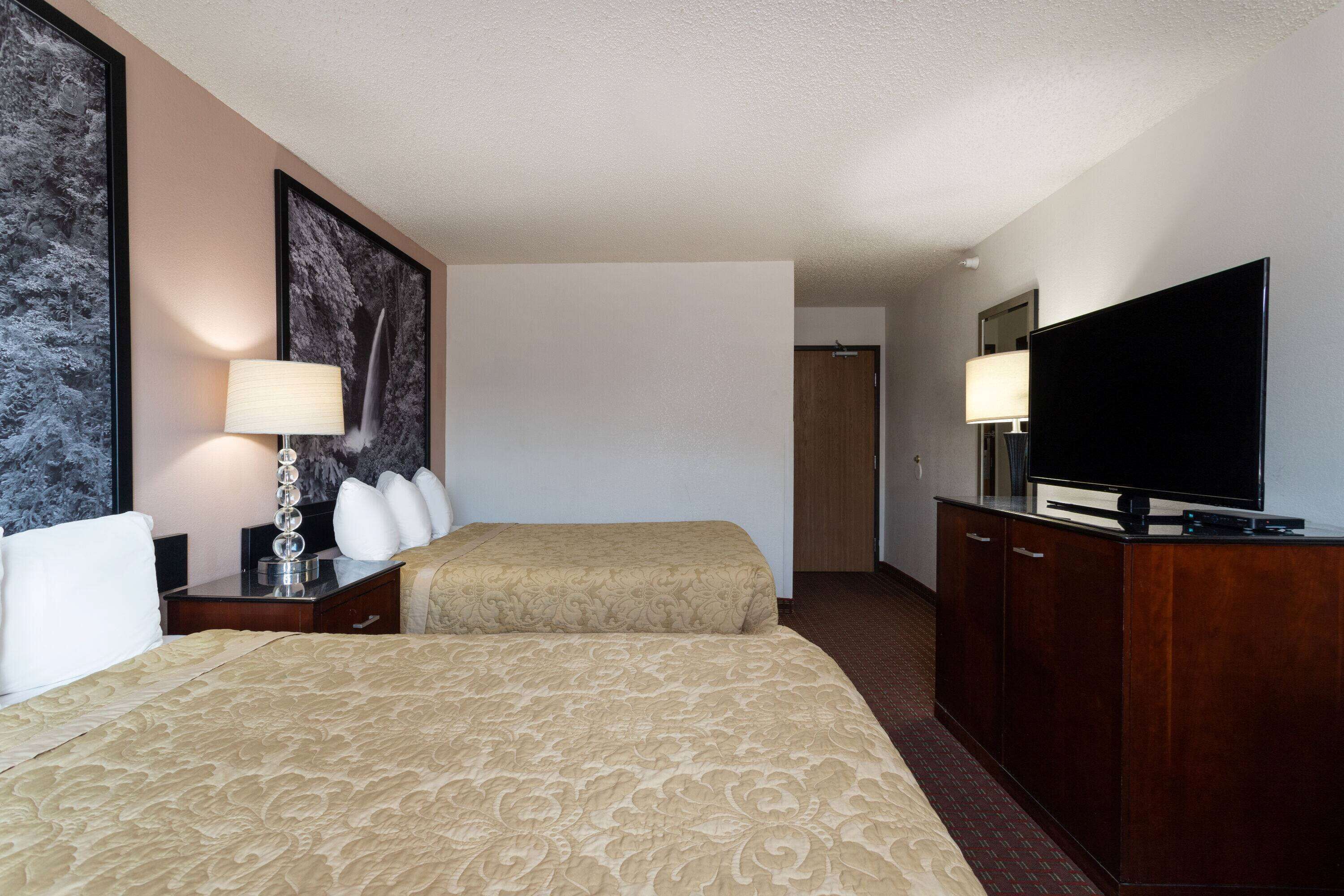 A hotel room with two beds, a TV, a desk with a lamp, and a large painting on the wall.
