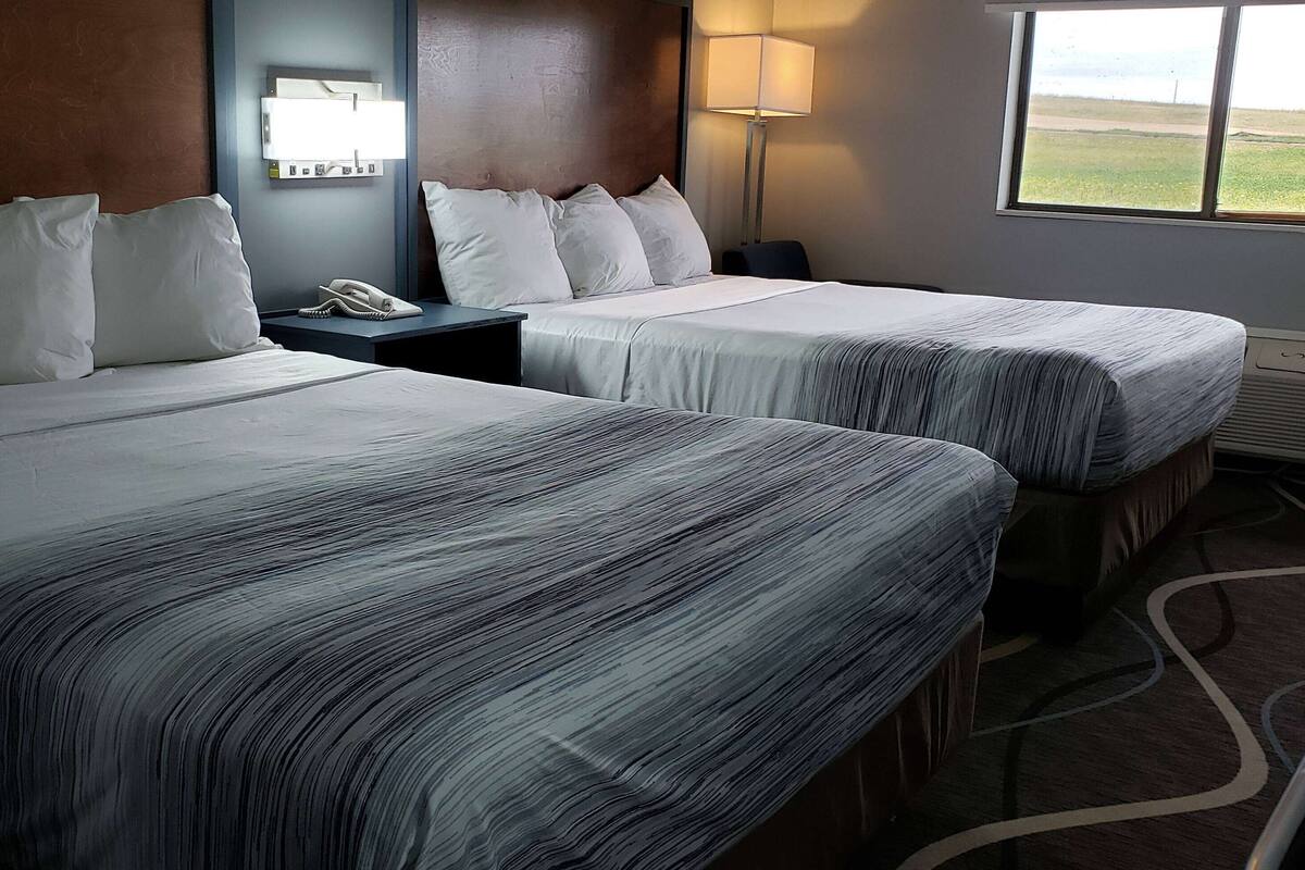 A hotel room with two beds, a nightstand with a phone, a lamp, and a window with a view of the beach.