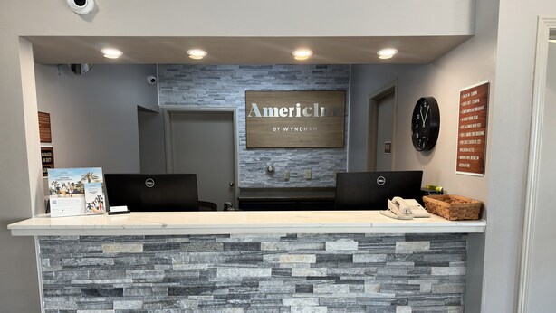 A reception desk with a stone facade, two Dell computers, a wooden sign above the desk, and a clock on the wall. - AmericInn by Wyndham Mauston (Mauston)