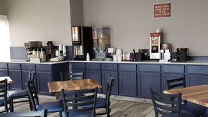 A café with wooden tables and blue chairs, a coffee machine, and a sign indicating breakfast hours. - AmericInn by Wyndham Mauston (Mauston)