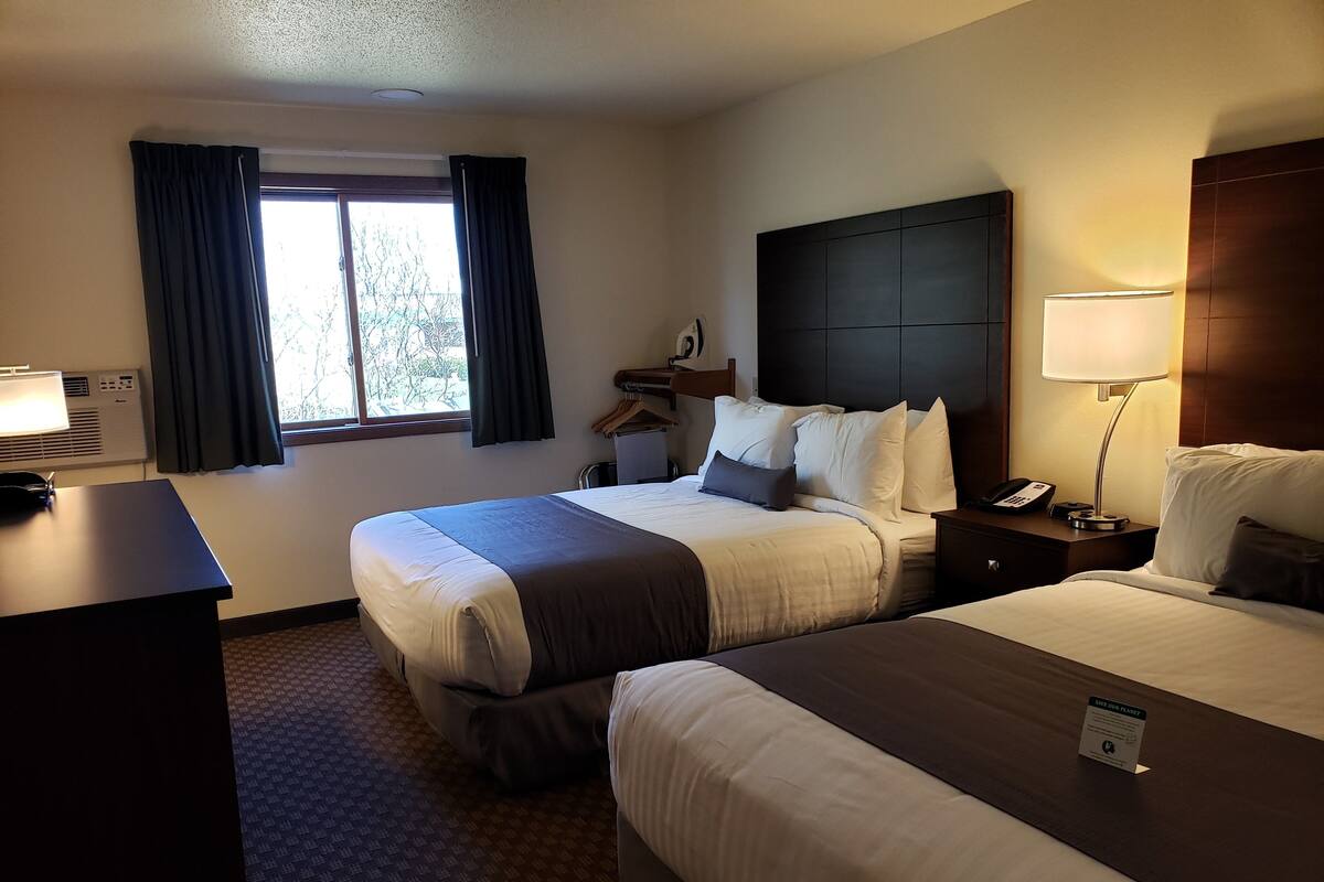 Quarto de hotel com duas camas, uma escrivaninha, uma luminária e uma janela com cortinas.