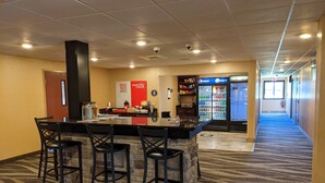A hotel lobby with a bar area, seating, a refrigerator stocked with beverages, and a carpeted floor.