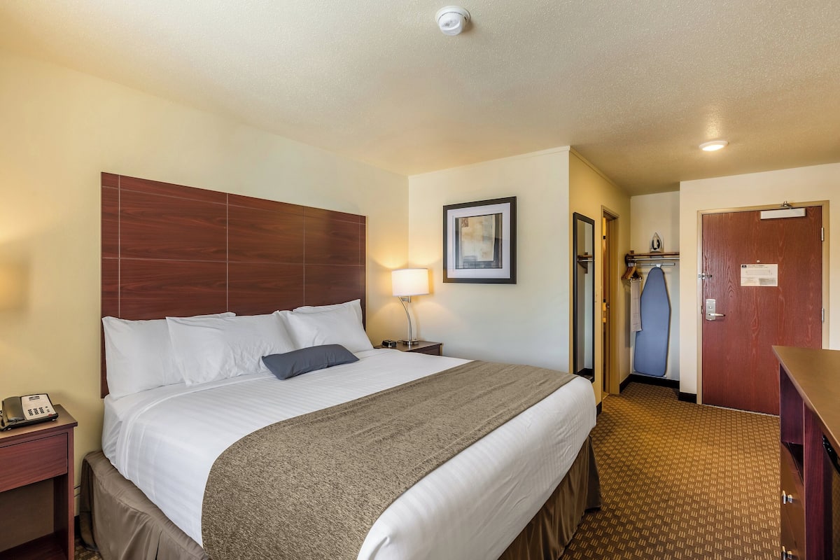 Quarto de hotel com uma cama grande, mesa de cabeceira com abajur, cabeceira de madeira, quadro na parede e porta com um aviso.
