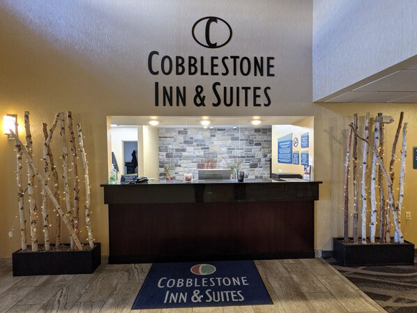 Un área de recepción con un cartel de Cobblestone Inn & Suites, un mostrador de recepción y ramas decorativas.