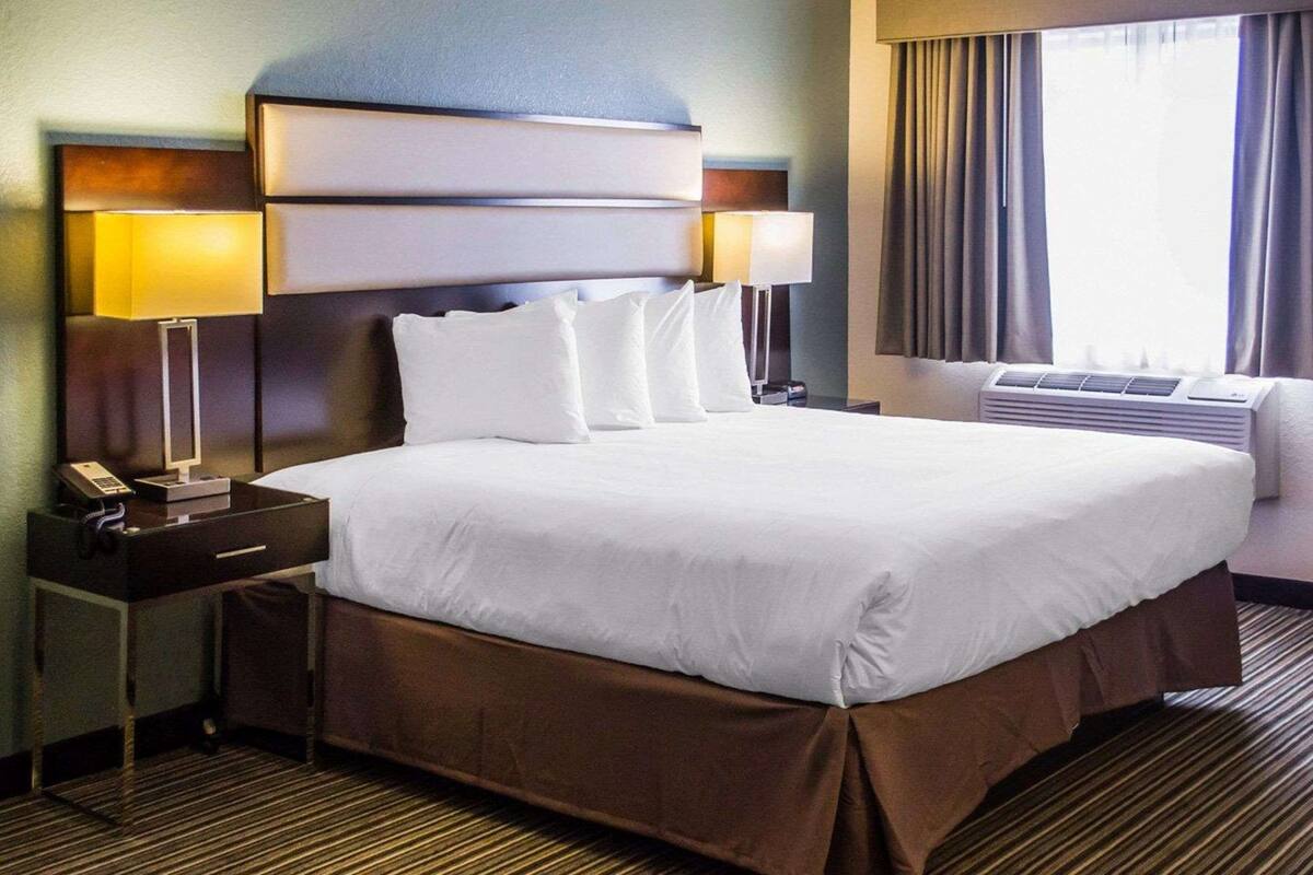 A hotel room with a large bed, a nightstand with a lamp, a window with curtains, and an air conditioning unit.