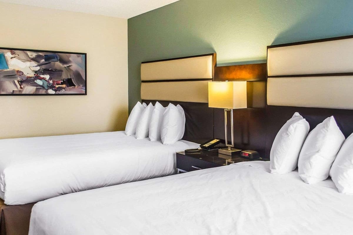 A hotel room with two beds, a nightstand with a lamp, a painting on the wall, and a clock.