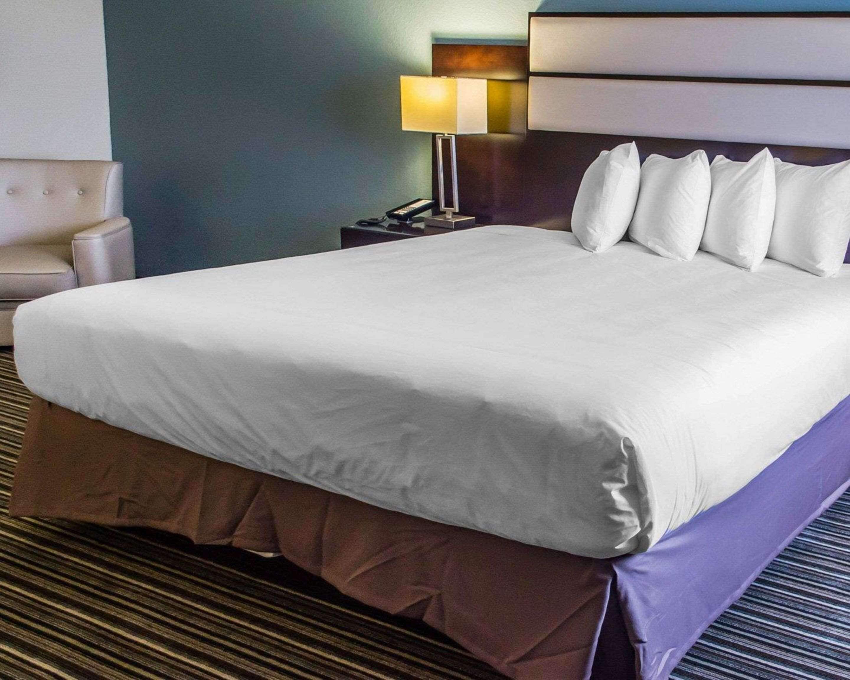 A neatly made bed with white linens and a brown skirt, a bedside lamp, and a chair in a hotel room.