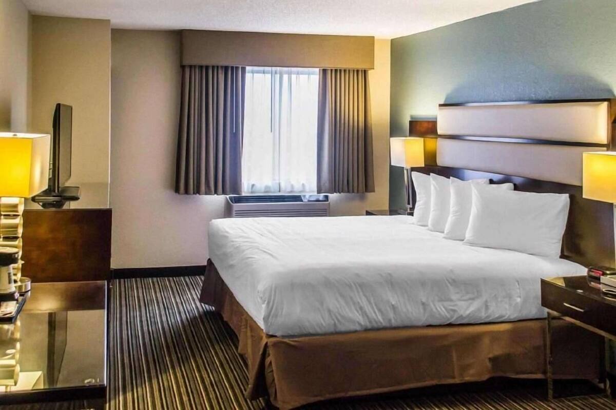 A hotel room with a bed, two bedside tables, a TV, a window with curtains, and a wall-mounted air conditioning unit.