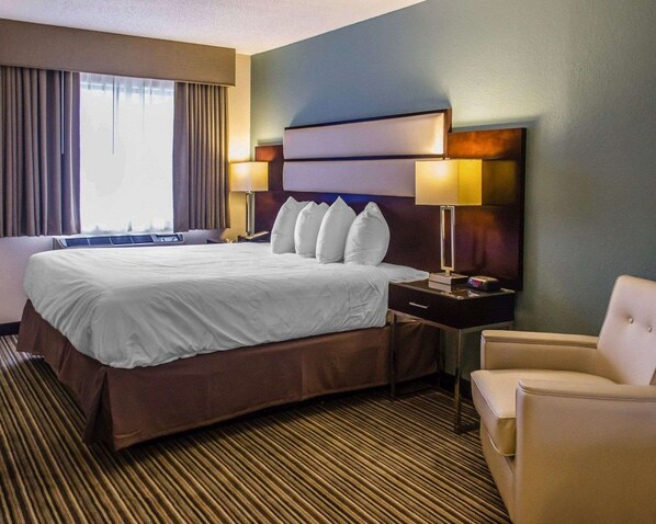 Habitación de hotel con una cama grande, una mesita de noche con lámpara, una silla y vistas al exterior a través de la ventana.