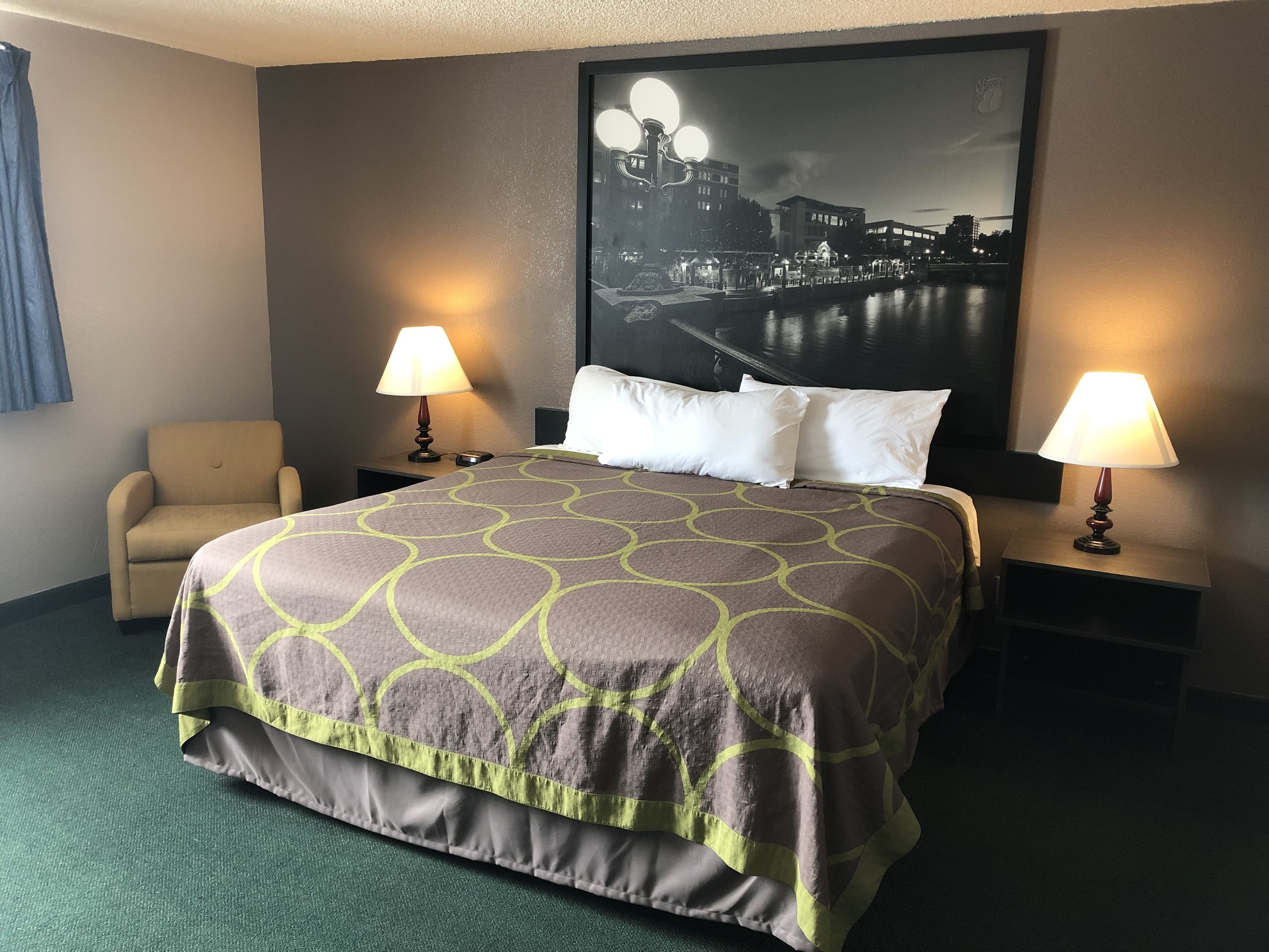 A hotel room with a large bed, two bedside lamps, a chair, and a cityscape painting on the wall.